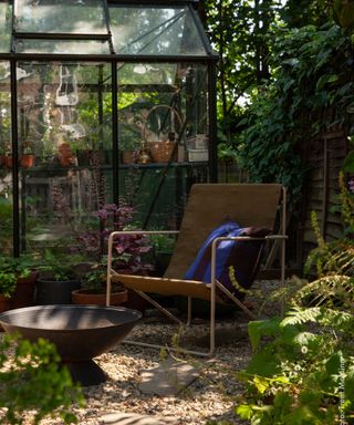 Secluded spot in garden with chair, fire pit and greenhouse, plant pots, flowers