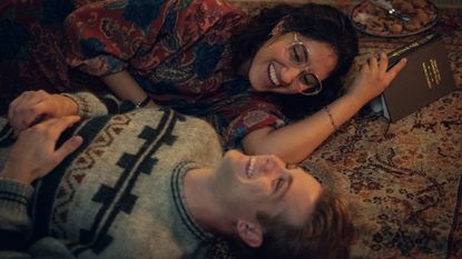 a woman (Ambika Mod as Emma) holds a book as she and a man (Leo Woodall as Dexter) lie on the floor while laughing