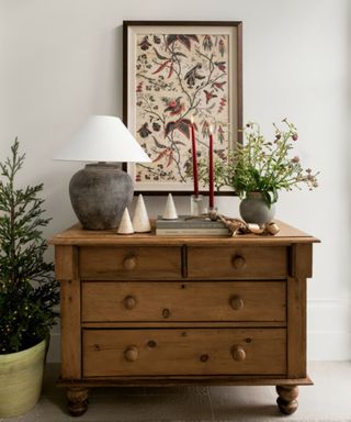 wooden chest of drawers with christmas decor, framed foliage print, lamp, red taper candles and vase of green foliage