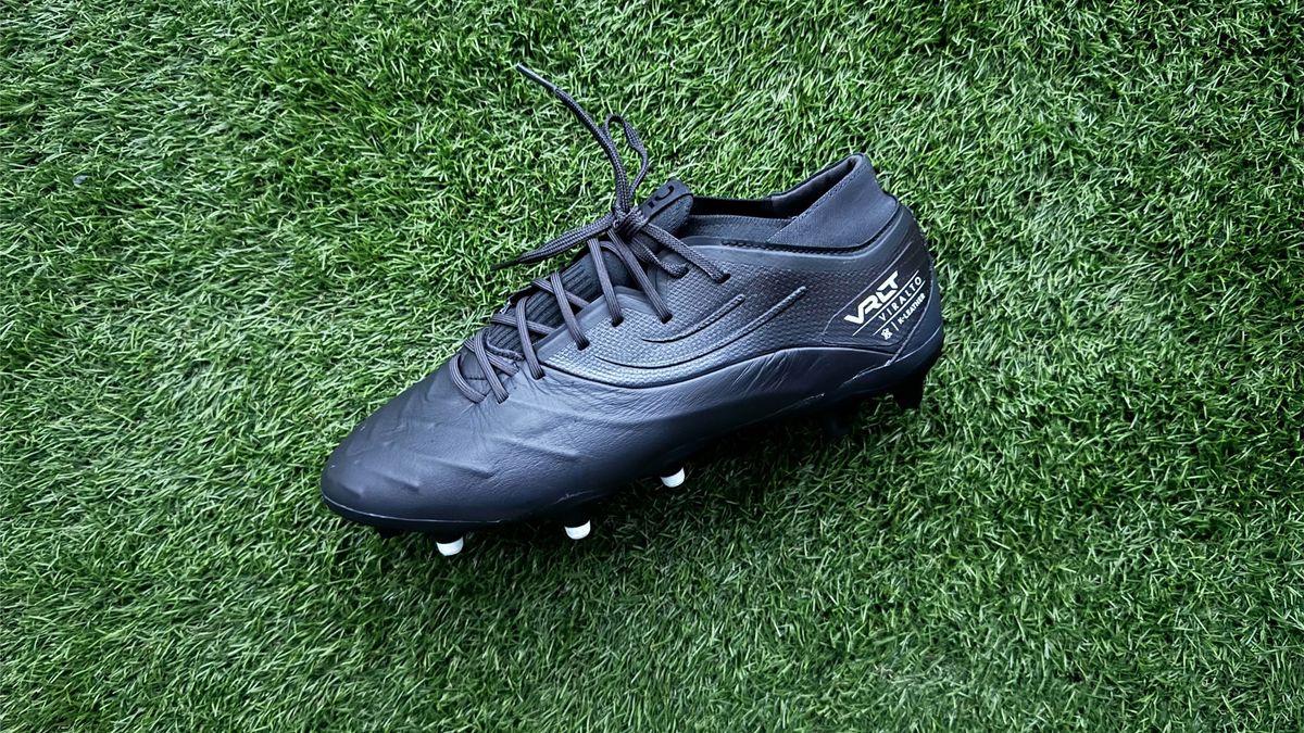 A black football boot with white studs is on some green artificial grass. Kipsta Viralto IV Pro