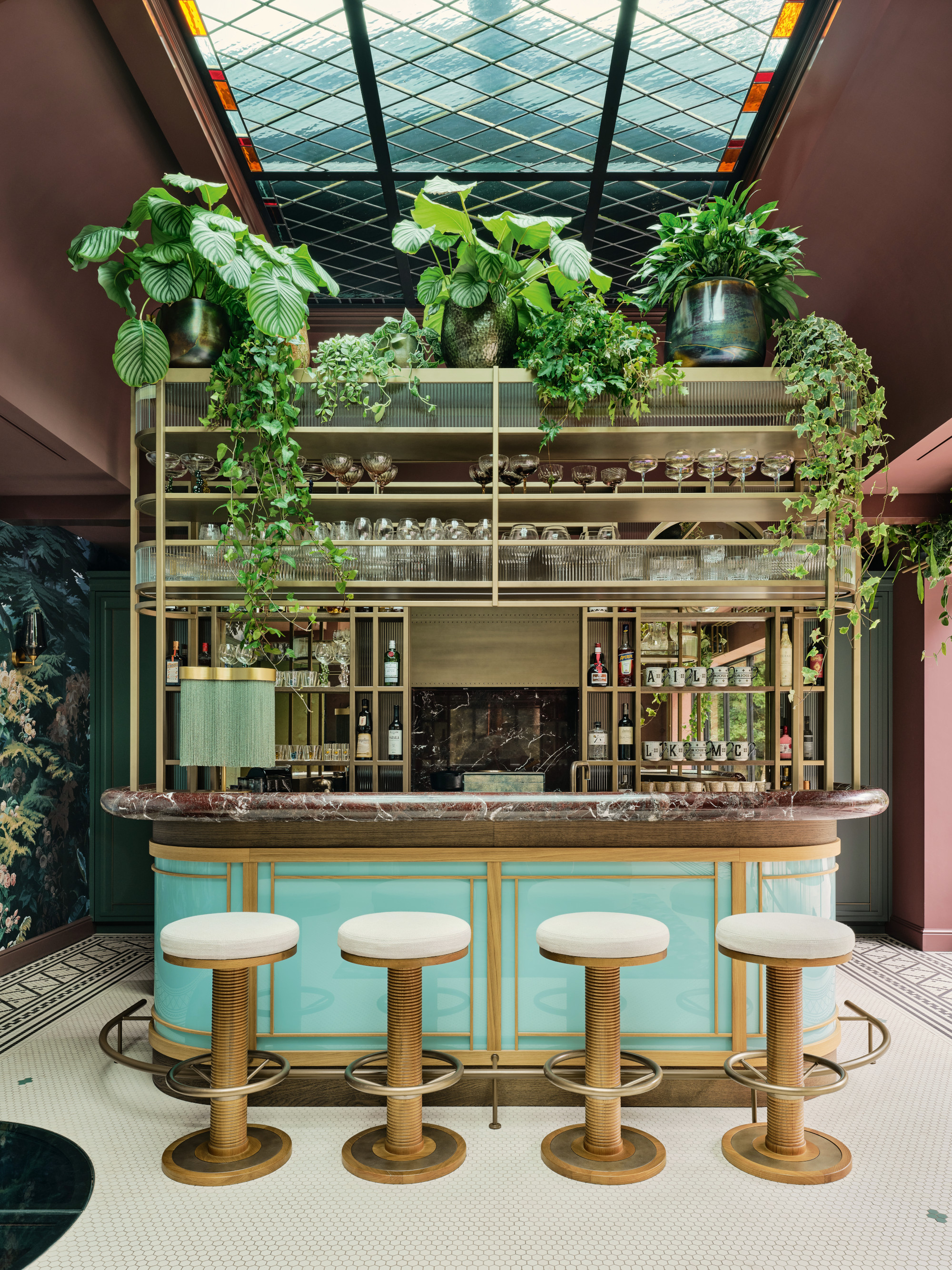 Home bar with blue body, burgundy marble countertop, brass and glass shelving and lots of trailing plants
