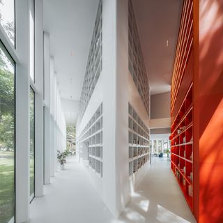 Narrow white floor and ceiling corridors, left hand side white shelving wall to ceiling unit, white planter, glass framed wall looking out to the external landscape, right hand side a white and an orange shelving floor to ceiling unit, blue seating area with plants and windows at the end of the corridor