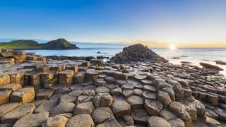 Giant’s Causeway at sunset, featuring interlocking basalt columns formed by ancient volcanic activity along the Northern Ireland coastline. The sun casts a golden glow over the hexagonal rock formations, with the ocean and green cliffs in the background.