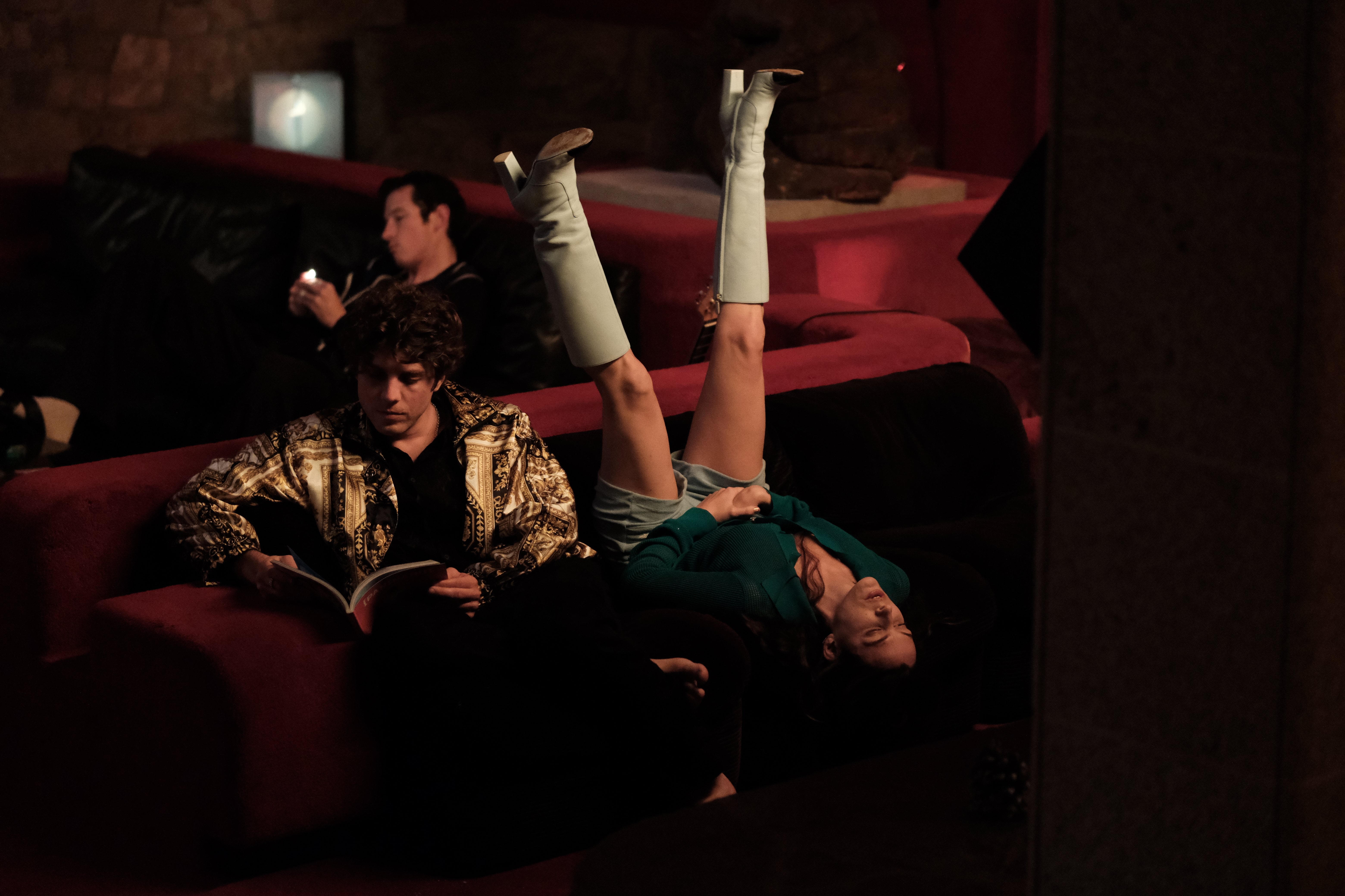 A young man in a golden jacket and a young woman in denim shorts, latex gray boots, and petrol green top, sitting upside down, relax in a living room, with another young man sitting behind them.