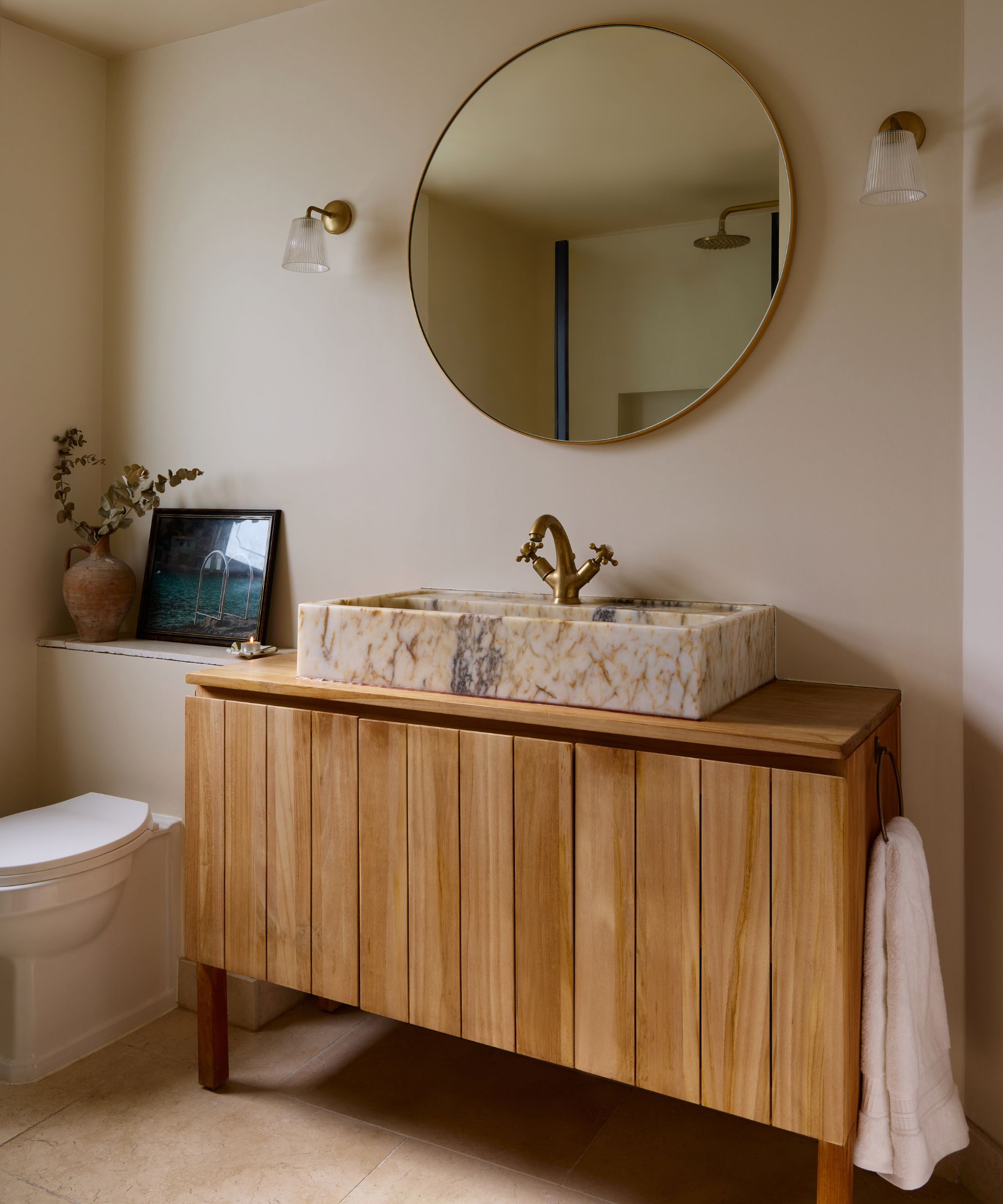 neutral modern cozy bathroom painted in Farrow &amp;amp; Ball's Dimity with a warm wood vanity and pink veined marble sink, round mirror, and brass wall lights