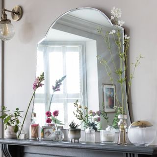 A mirror rested on the mantel of a fireplace surrounded by bud vases with flowers and a wall light above