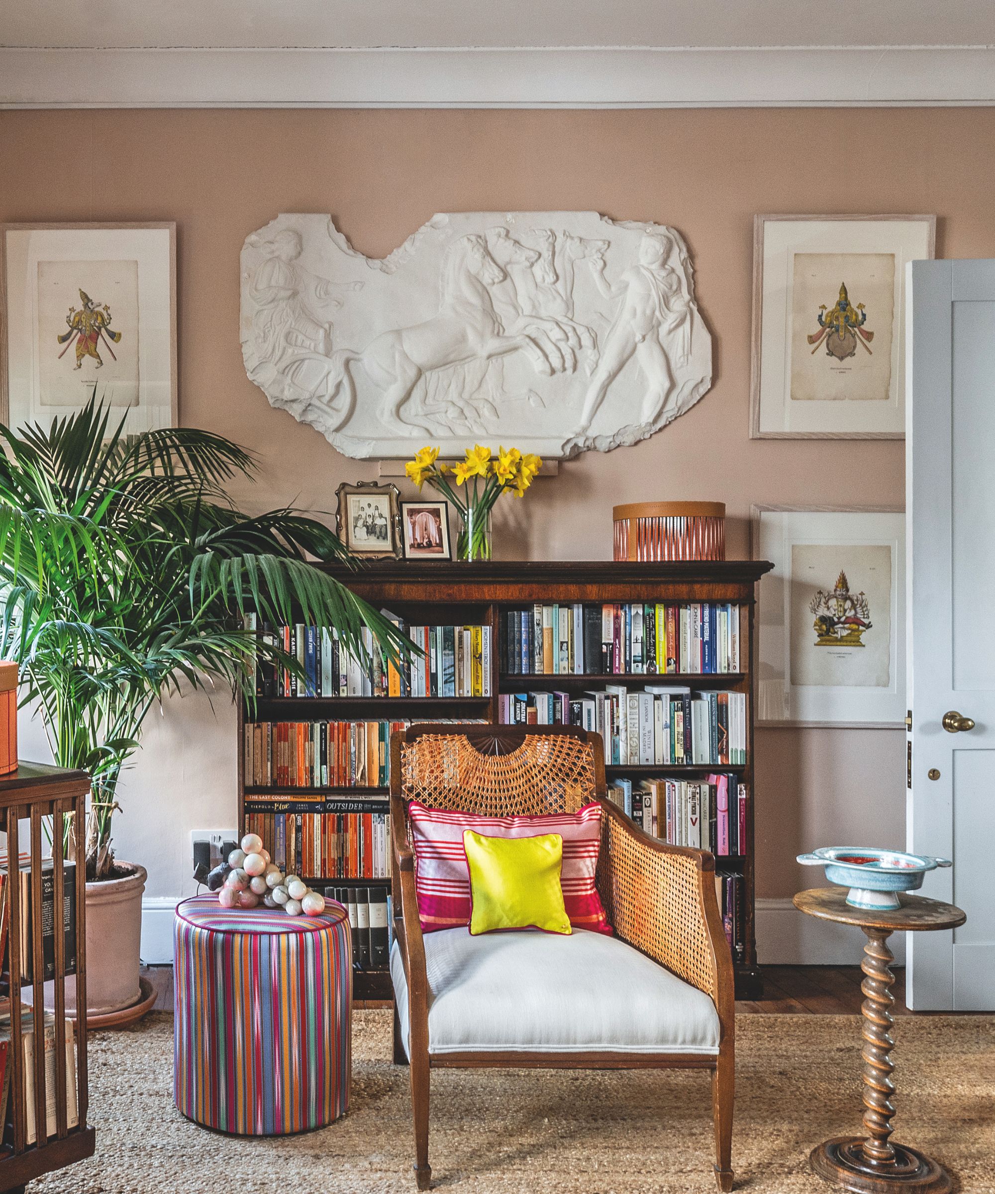 A cozy reading nook with a wooden bookshelf, a cane-back chair with a bright yellow pillow, and a large classical plaster relief sculpture mounted on a pink wall