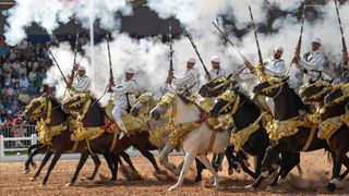 A line of horsemen performs T'bourida (Fantasia) during the Horse Show of El Jadida, while firing muskets, creating rising smoke in the air 