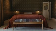 A bedroom with patterned wallpaper, wall sconces, a striped bolster pillow, a trailing cinnamon-colored duvet, and an end of bed bench