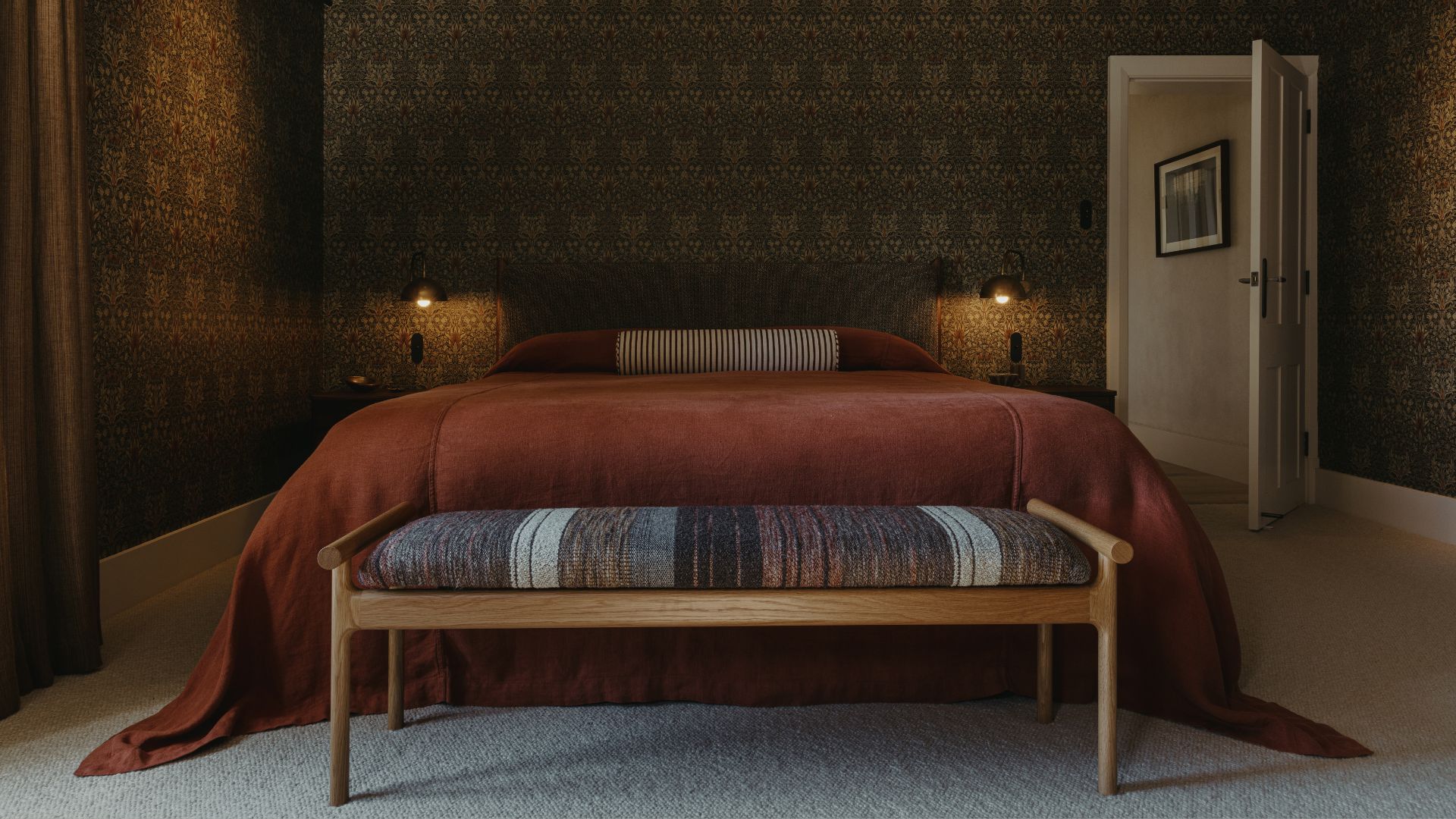 A bedroom with patterned wallpaper, wall sconces, a striped bolster pillow, a trailing cinnamon-colored duvet, and an end of bed bench