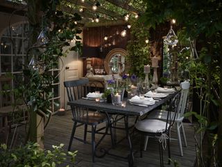 A beautiful indoor-outdoor pergola, with a 6-seater dining table and chairs, and lots of lit candles, twinkling fairy lights, and climbing plants, and a white door to the left