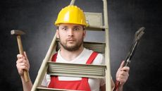 A man wearing a hard hat has tools in hand, a ladder over his head and a perplexed look on his face.