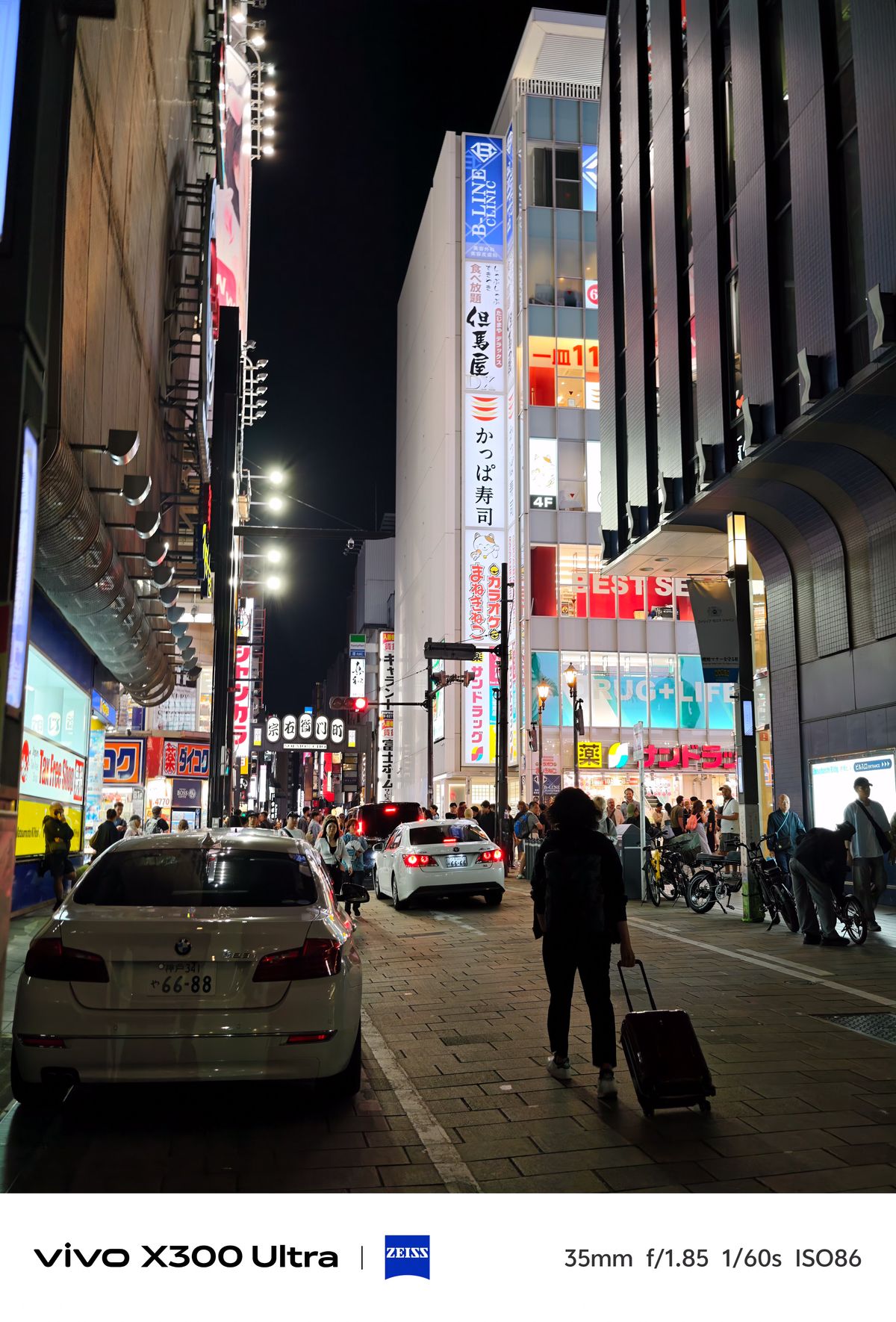 A night street scene with cars, pedestrians, and a person rolling a suitcase past bright shop facades.