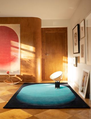 A rug with a blue oval on black background placed on the parquet floor of a room featuring wooden walls with a further rug in white and red hanging vertically