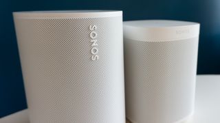 Sonos Era 100 i hvidt på et hvidt bord og blå baggrund ved siden af Sonos One