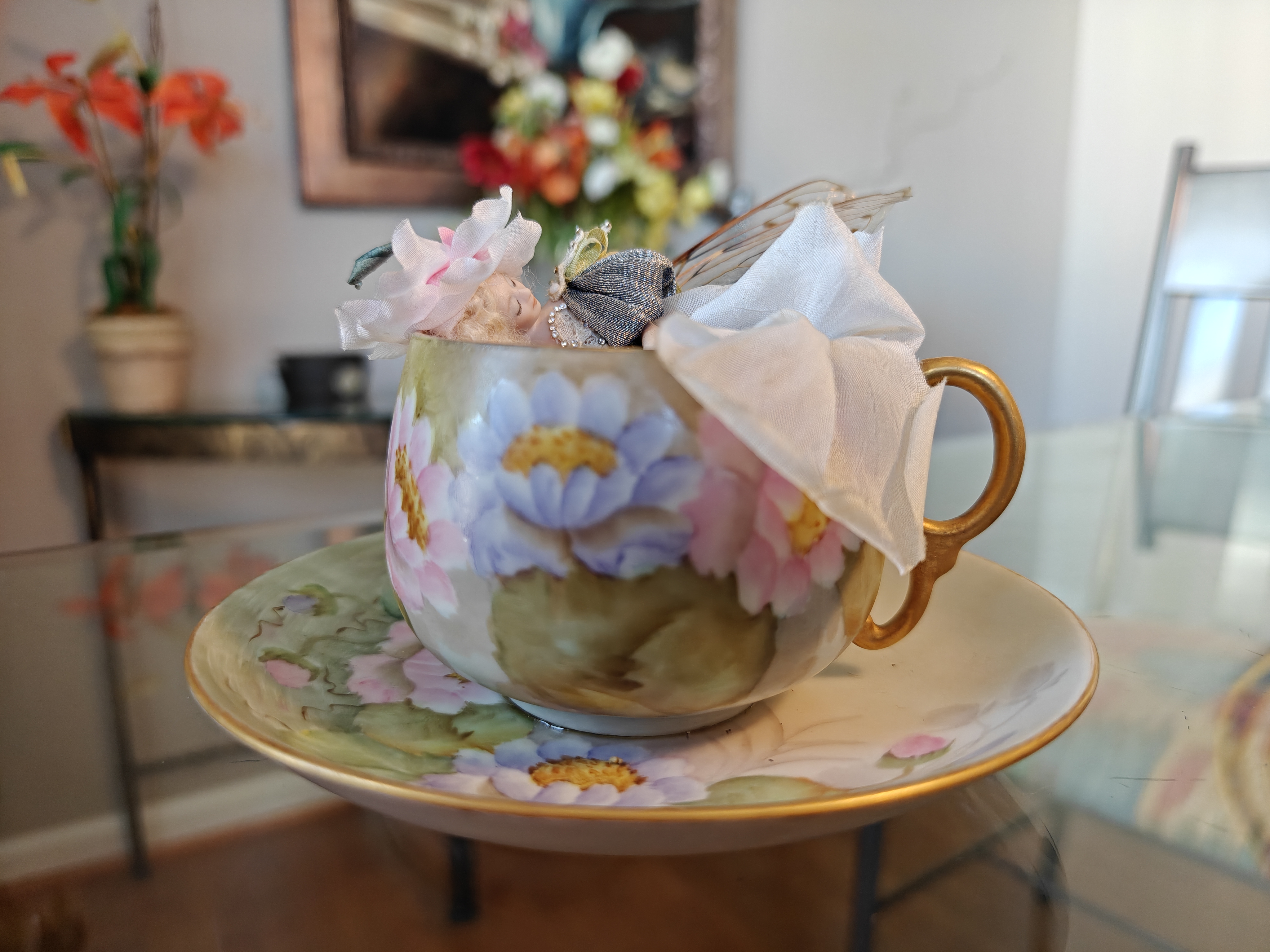 A teacup fairy with insect wings on a glass tabletop. She is asleep and wearing a fancy dress.