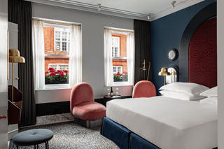 The chubby furniture and playfully beautiful interiors of an hotel room feature coral pink upholstered velvety armchairs, golden standing lamps, a chunky bed, and striking panel screens.