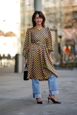 A guest wears a navy blue / pink / green / yellow print pattern long sleeves / belted knees dress, a black shiny leather handbag, blue faded denim rolled-up pants, black shiny leather pointed pumps heels shoes , outside Kate Spade, during New York Fashion Week, on February 10, 2023 in New York City.