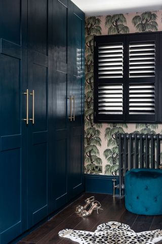 A dark blue wardrobe with black shutters in bedroom and leopard print rug, heeled shoes, palm print wallpaper, grey radiator and teal footstool