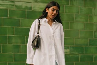 Woman wearing Madewell button-down white shirt, light wash jeans, and black shoulder bag stands in front of green wall.