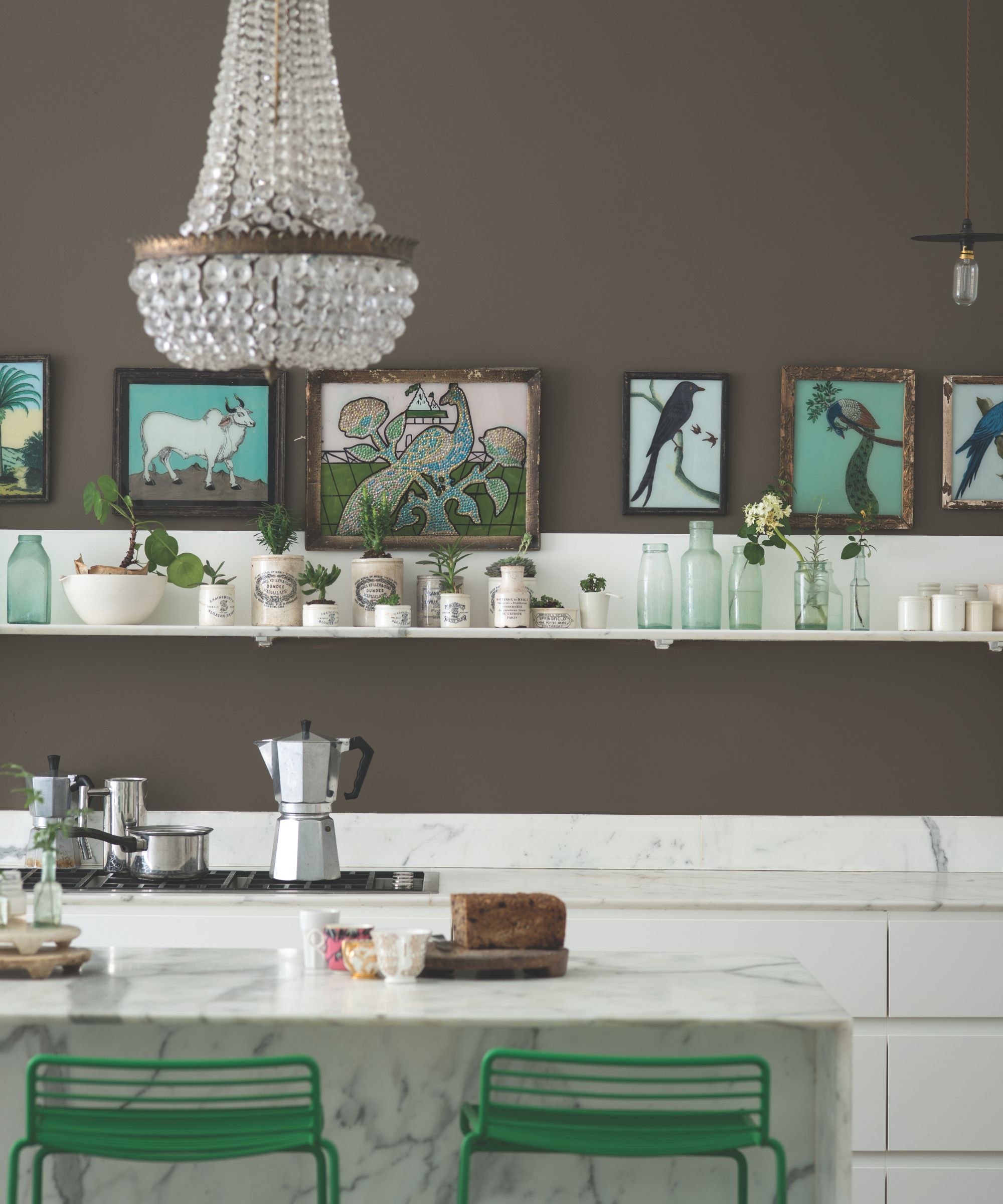 Kitchen with chandelier at the centre, a marble island, green chairs tucked underneath, marble countertops and hob with mokka pot, shelf with vintage glass bottles and ceramic pots, and a dark brown painted wall with bird prints along it
