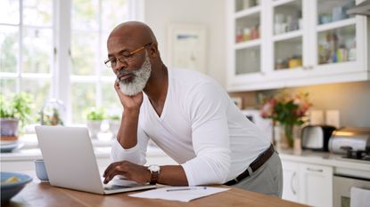 An older man leans on his kitchen counter as he looks at his laptop.