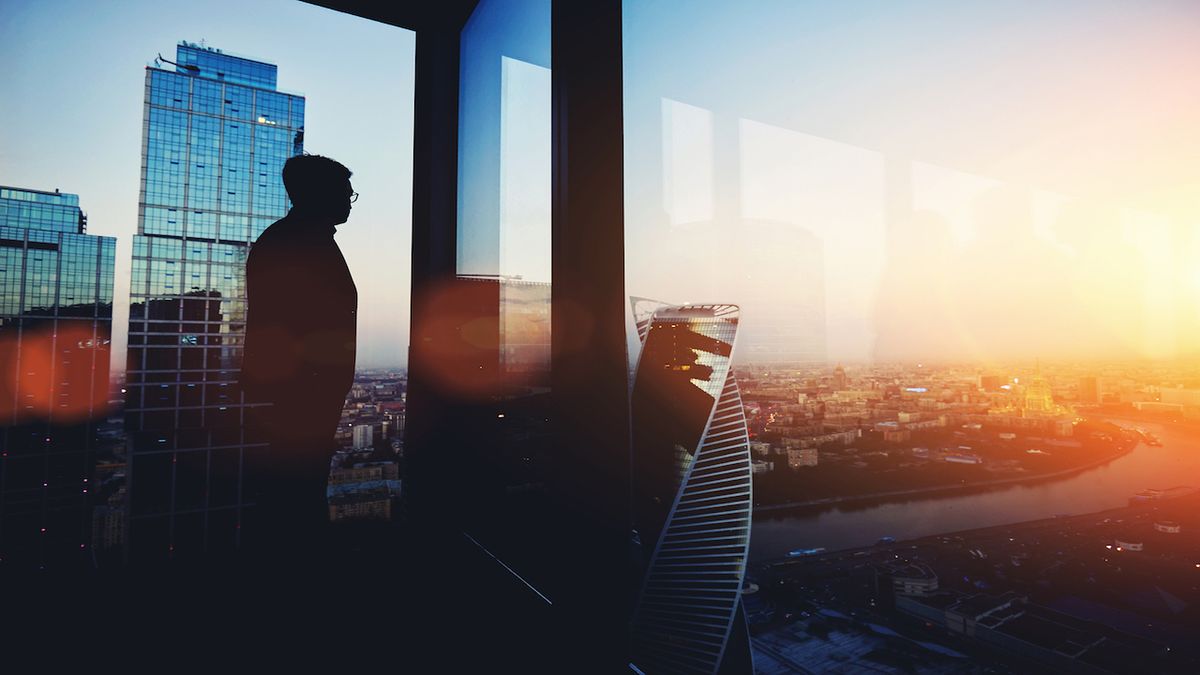 A silhouette of a figure observing a city at sunrise from an office window