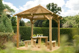 Wooden slat roof gazebo with outdoor table and benches sits within surrounding hedging