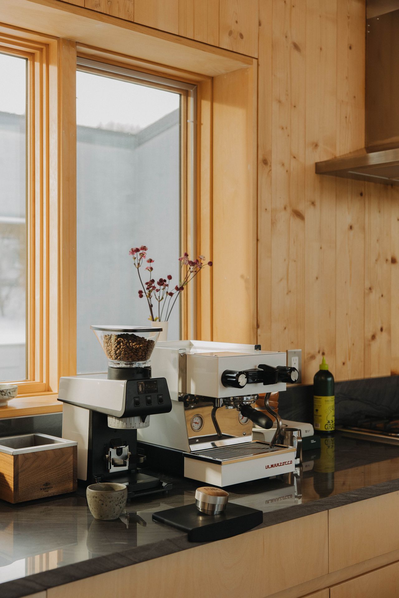 A white La Marzocco machine next to a matching La Marzocco grinder on a black countertop in a timber kitchen.