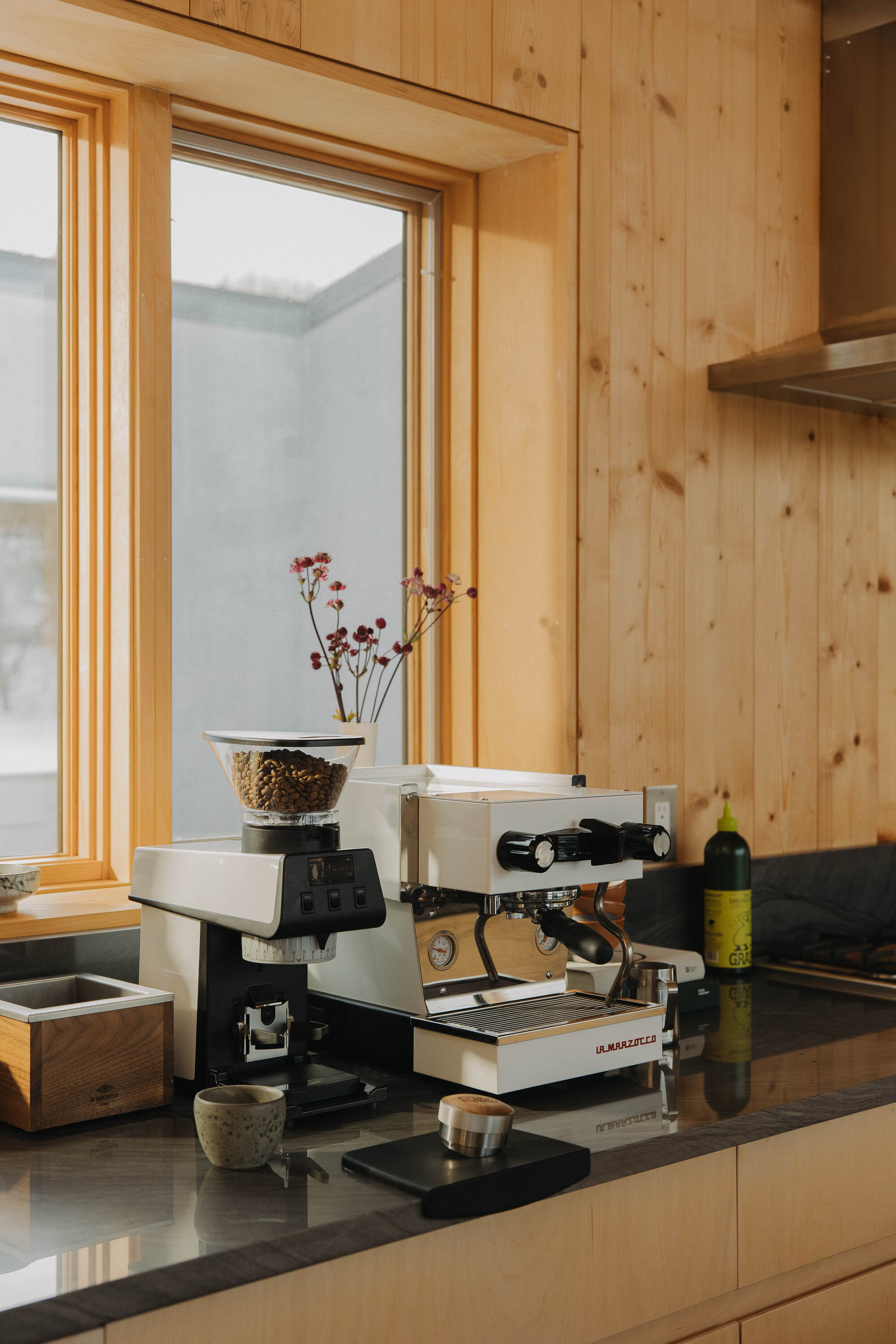 A white La Marzocco machine next to a matching La Marzocco grinder on a black countertop in a timber kitchen.
