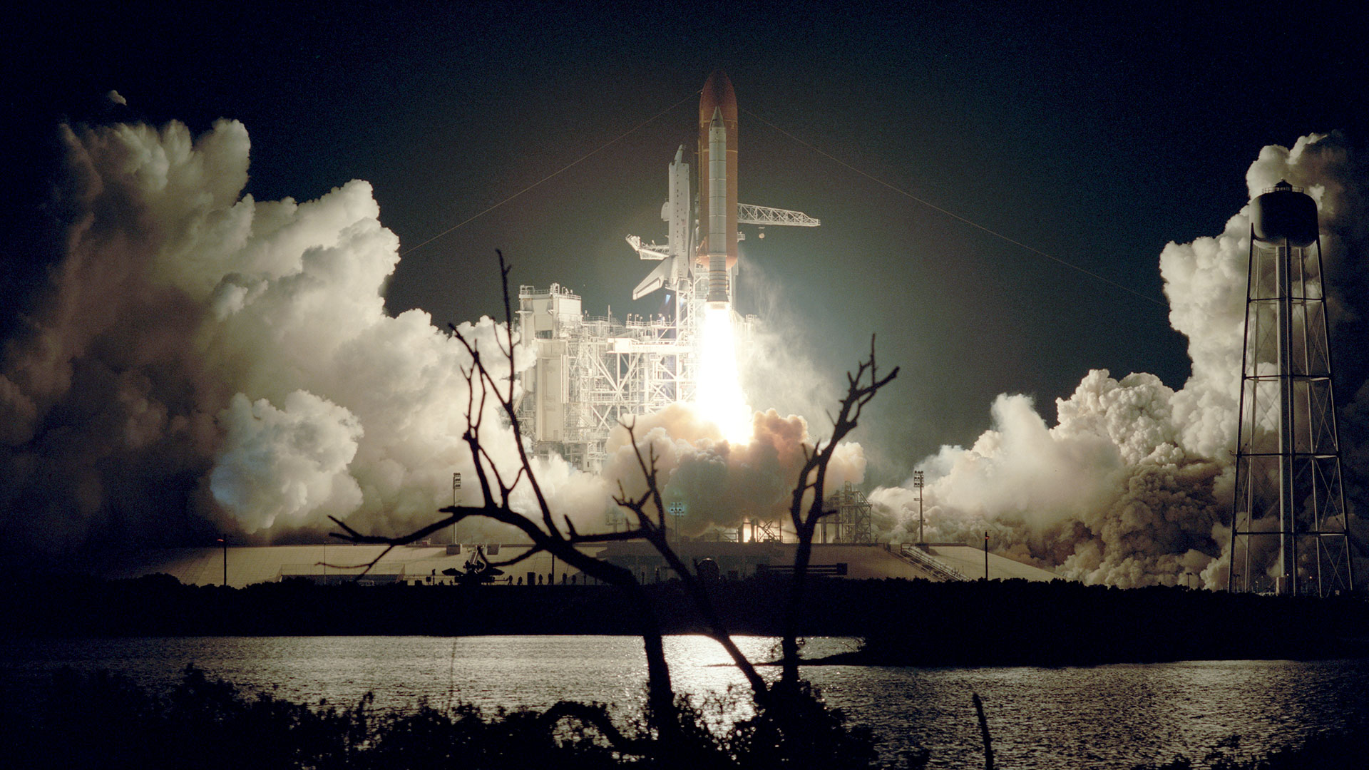 a winged orbiter attached to twin rockets and an orange-brown fuel tank lifts off at night, its plume lighting its launch pad and the water below.