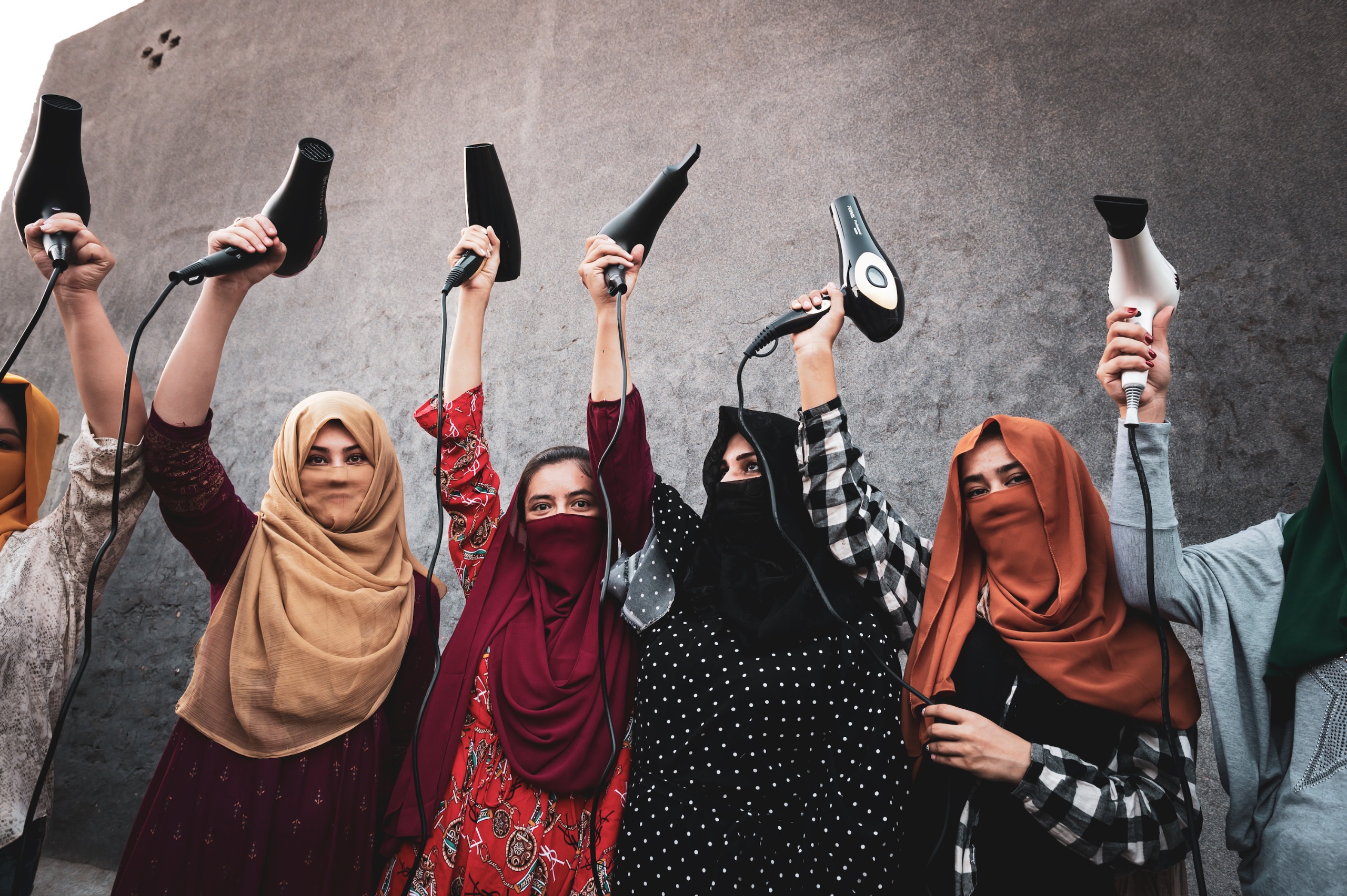 A row of Afghan refugee women in brightly coloured headscarves, most with their faces veiled, stand against a grey concrete wall holding hairdryers triumphantly above their heads, cables dangling.