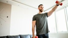 man wearing a grey tshirt raising one arm in front holding a small red dumbbell. there's a plain wall and a window behind him in a living room setting.
