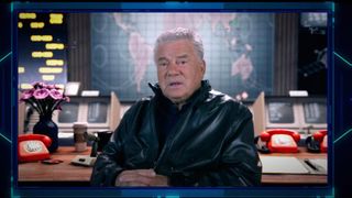 a man in a black leather jacket sits at a desk with a map of the world in the background.