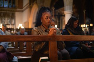 a woman (Dionne Brown as Queenie) prays in a church among other parishioners, in 'Queenie'