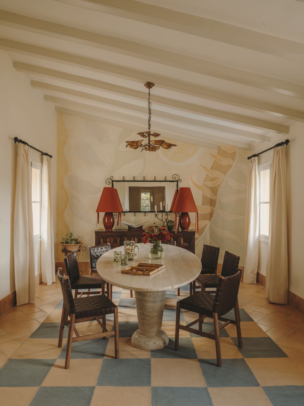 A murals-clad livingroom with wooden floors is brought to life by a checkered cream and pale blue rug, a sculptural marble table, and two matching red lamps.