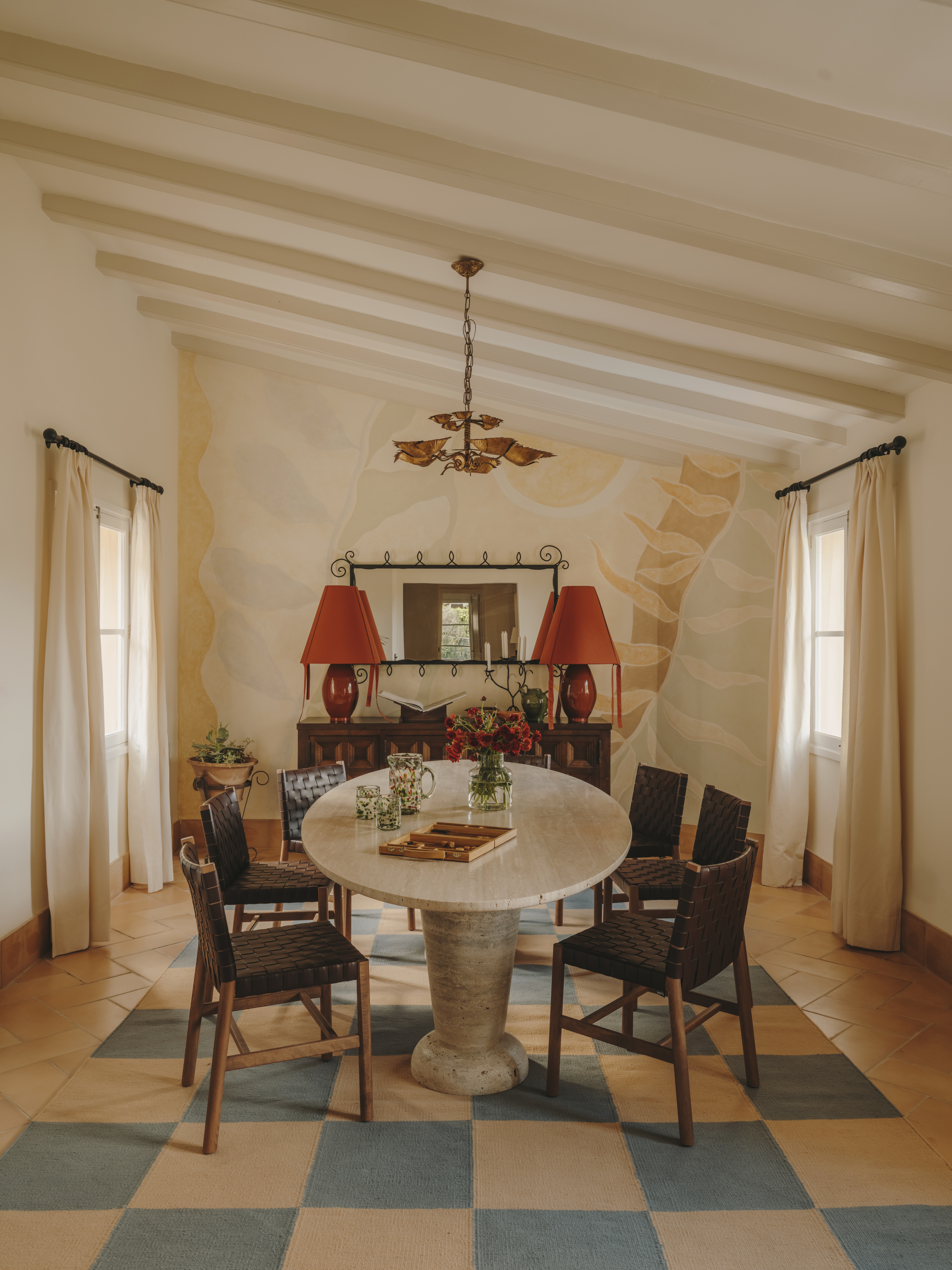 A murals-clad livingroom with wooden floors is brought to life by a checkered cream and pale blue rug, a sculptural marble table, and two matching red lamps.
