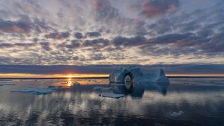 a serene landscape with calm water below and a distant setting sun on the horizon with large icebergs in the water below.