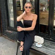 Influencer wears metal detail sandals with black trousers and a black strapless top. She sits on a bench outside of a coffee shop.