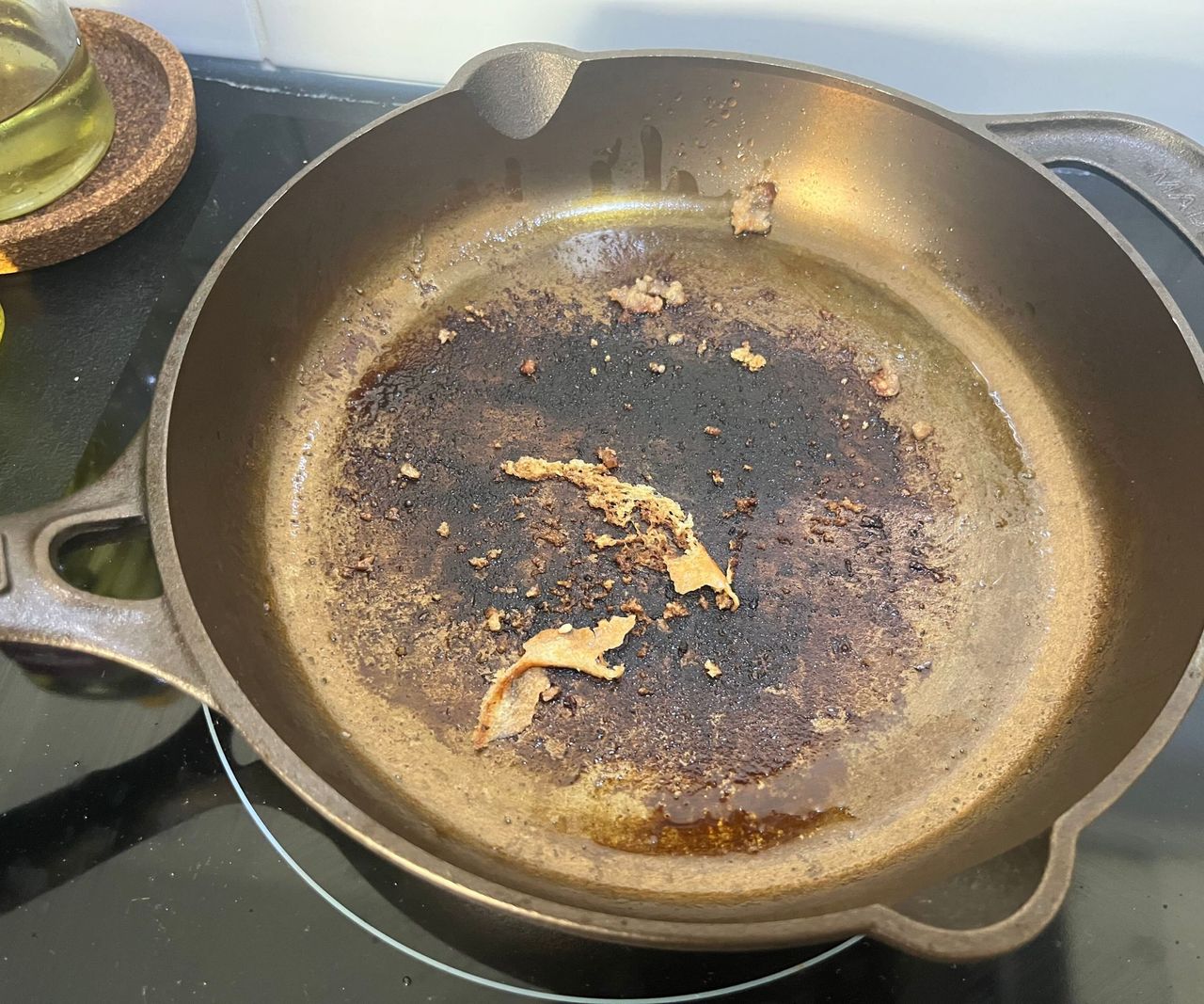 A small bronze colored cast-iron skillet on a black glass stove with burn marks inside.