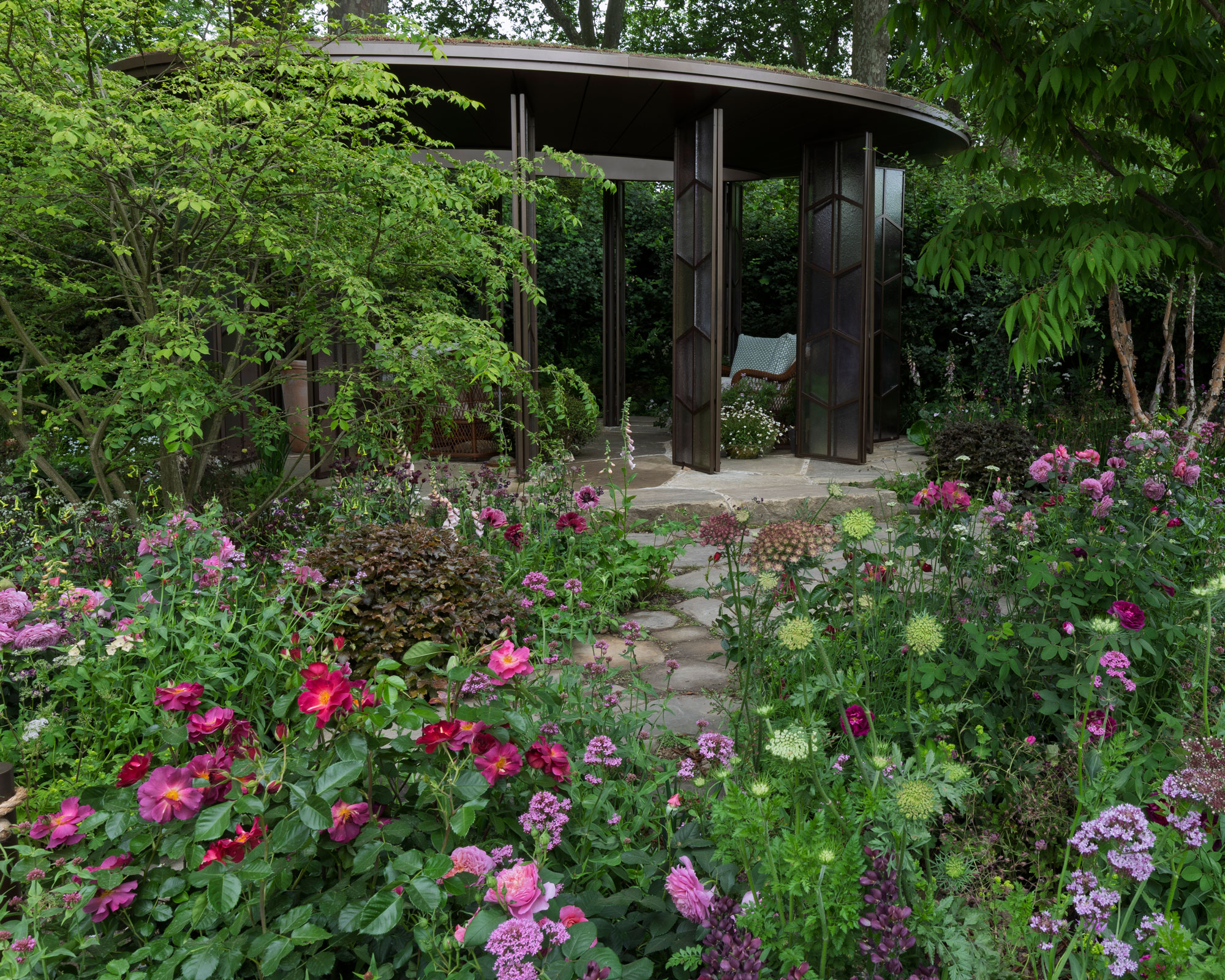 garden pavilion with garden chair, surrounded by trees and flowerbeds, with a crazy paving path