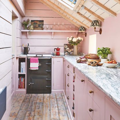 pink painted galley kitchen with small black stove, pink Shaker-style cabnets, open shelves, skylights, and wood panelling