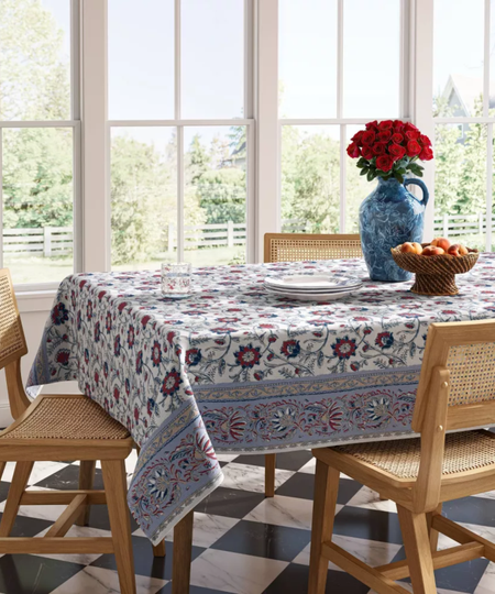 a dining table with a red and blue floral table cloth, a vase with red roses, and light wood dining chairs