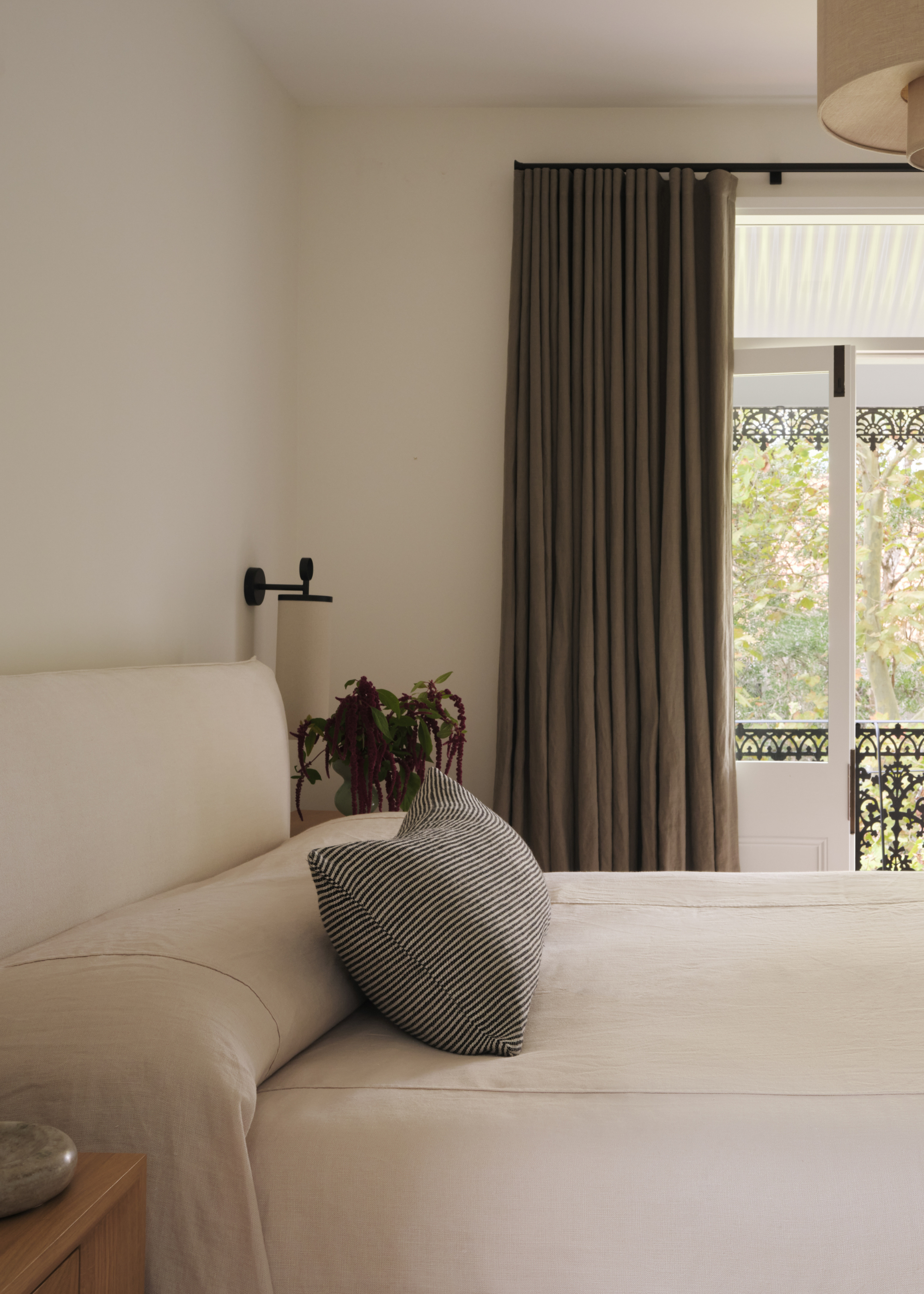 A white bedroom with brown window treatments leading out to a balcony, a white bedscape, a striped throw pillow, a vase with a floral arrangement, and a black wall sconce