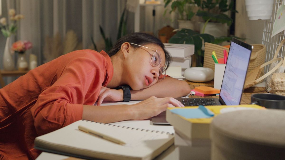 young woman sits at a desk surrounded by books and a laptop. She has her head laid down on the desk as she reads something on the laptop