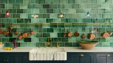 A green tiled kitchen with marble countertop and blue cabinets, with copper pans hanging from a rod and colorful glass pendant lights