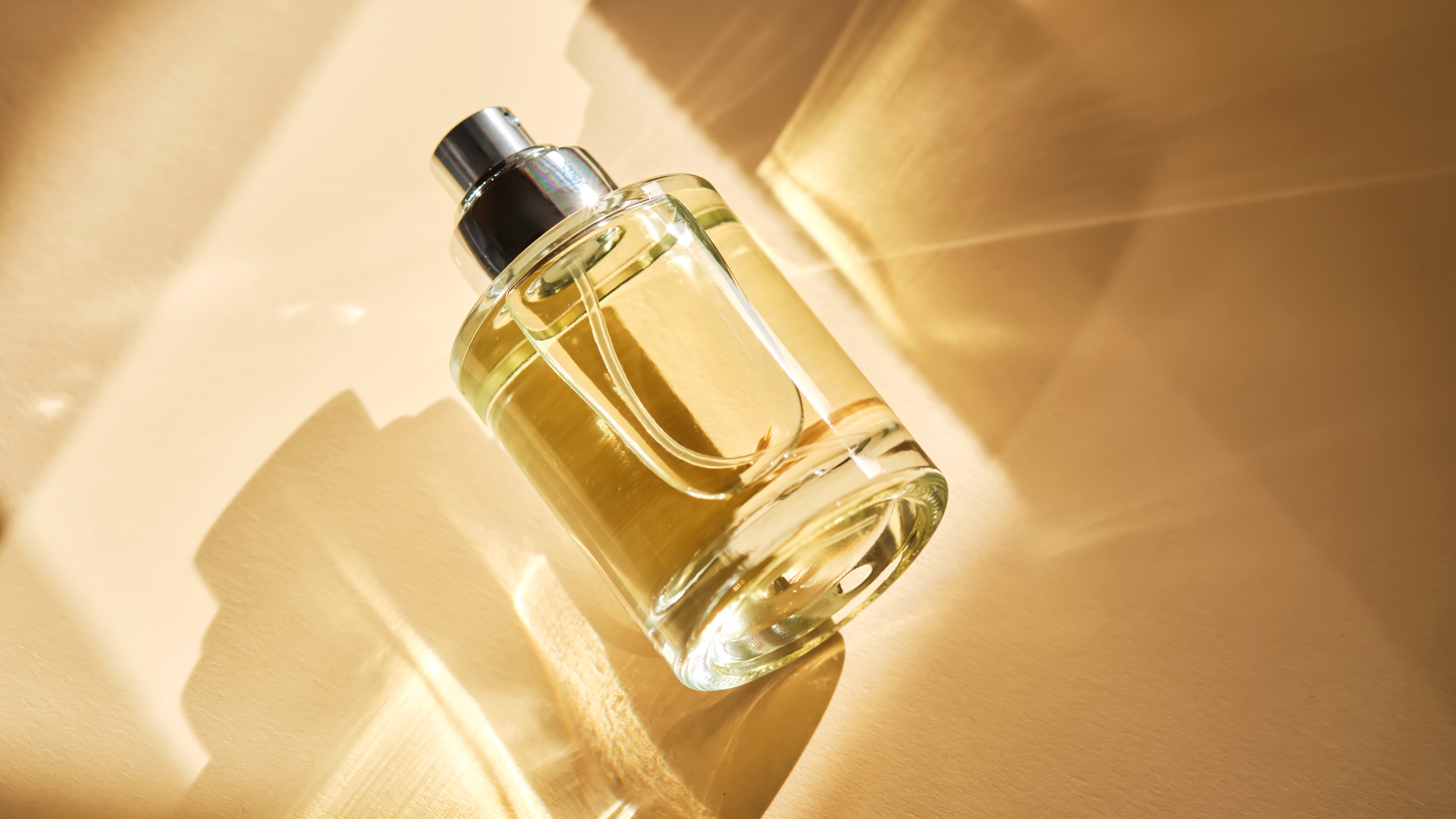 A perfume bottle with light reflecting through it against a cream background and surrounded by shadows