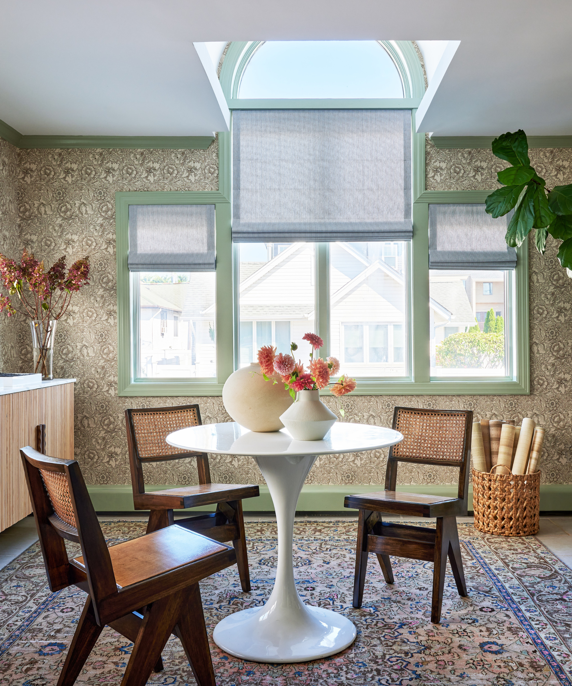 A light-filled dining room with sage green trim, floral wallpaper, and a small round dining table surrounded by three wooden chairs