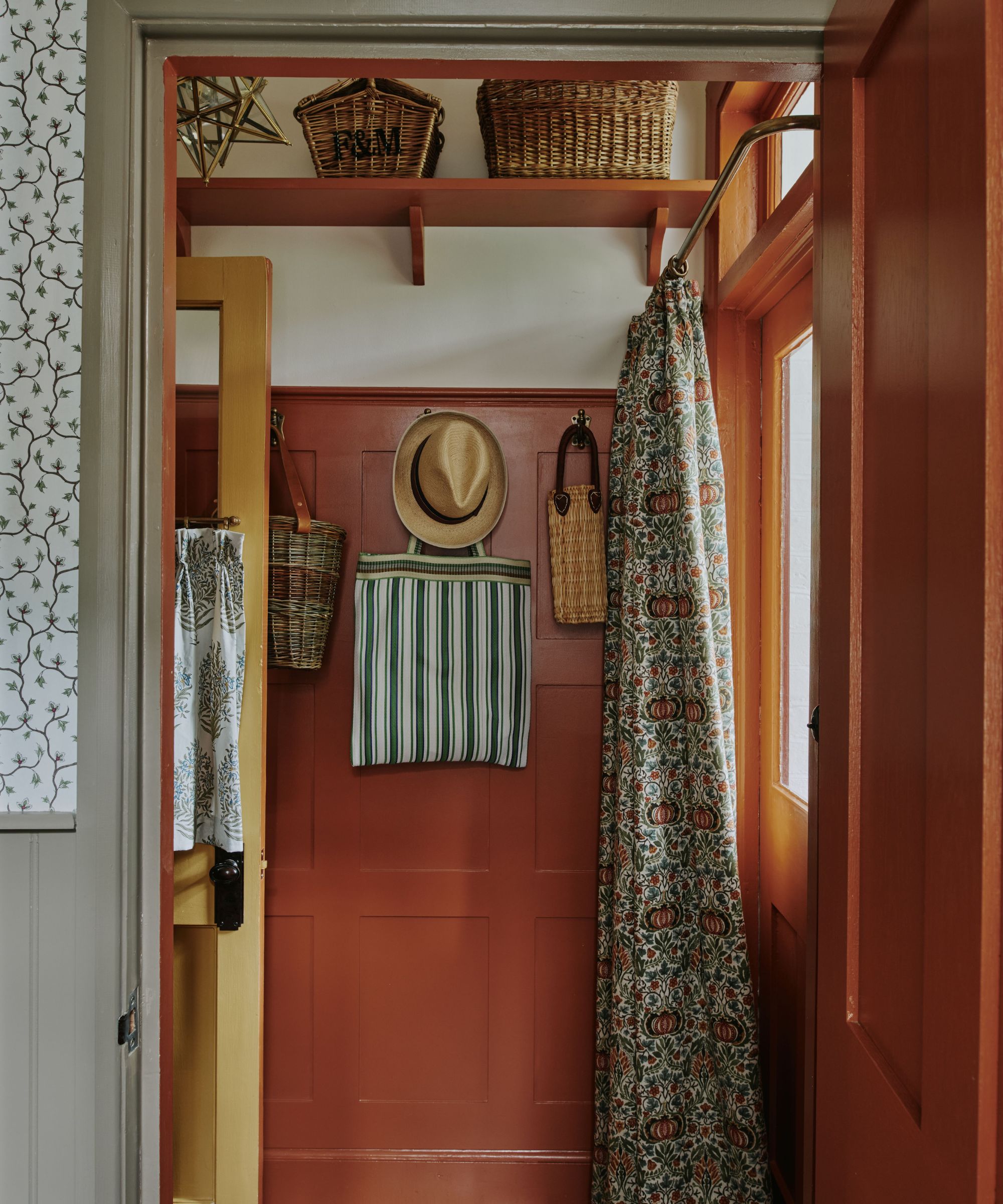 Entryway with floral curtains, red painted walls and panelling, hooks hanging bags and hats, and a shelf of baskets on the top