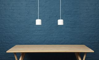 Blue painted brick wall backdrop, long light wood table, two white ceiling lights suspended above the table
